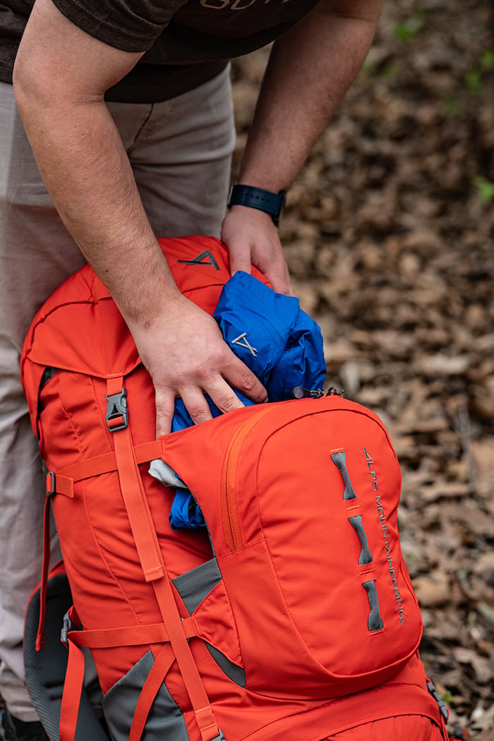 Hiker placing gear into the front kangaroo pocket of a Chili Red Tail 65L pack