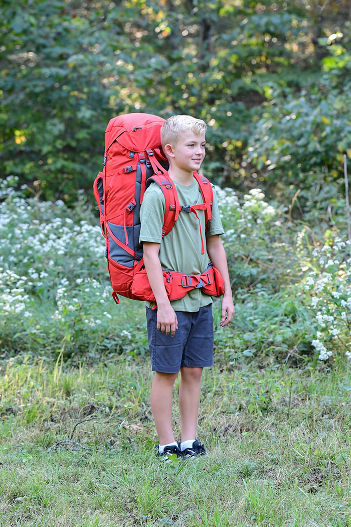 Hiker wearing a Chili Red Tail 65L pack, viewed from the front on a wooded trail