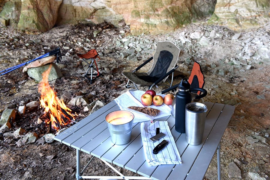 Silver ALPS Camp Table holding dishes, fruit and cookware beside a campfire