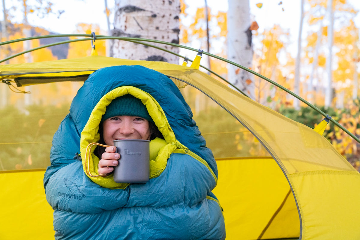Camper holding a mug while sitting in the hooded Peregrine Anatum sleeping bag