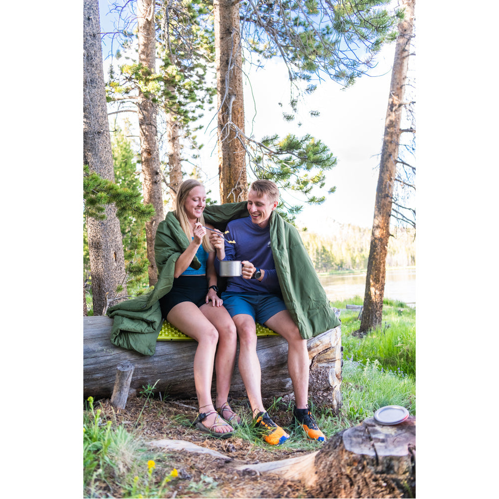 Two campers sharing an olive Peregrine Link-Up field quilt on a log